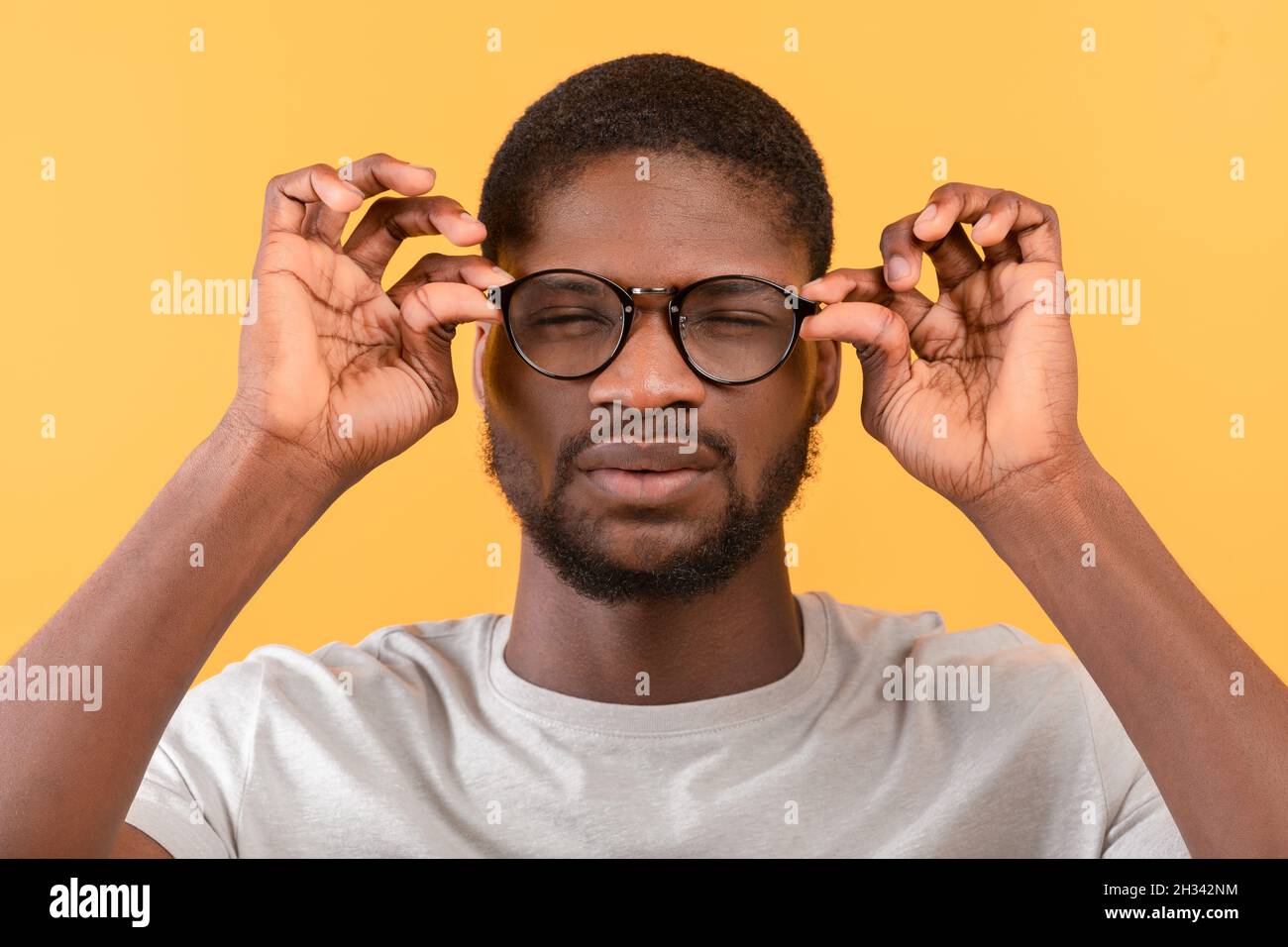 afroamerikanischer Mann mit schlechter Sehkraft, der eine Brille trägt und Augen schielt, während er durch eine Brille auf die Kamera blickt, gelber Studiohintergrund. Gesund Stockfoto