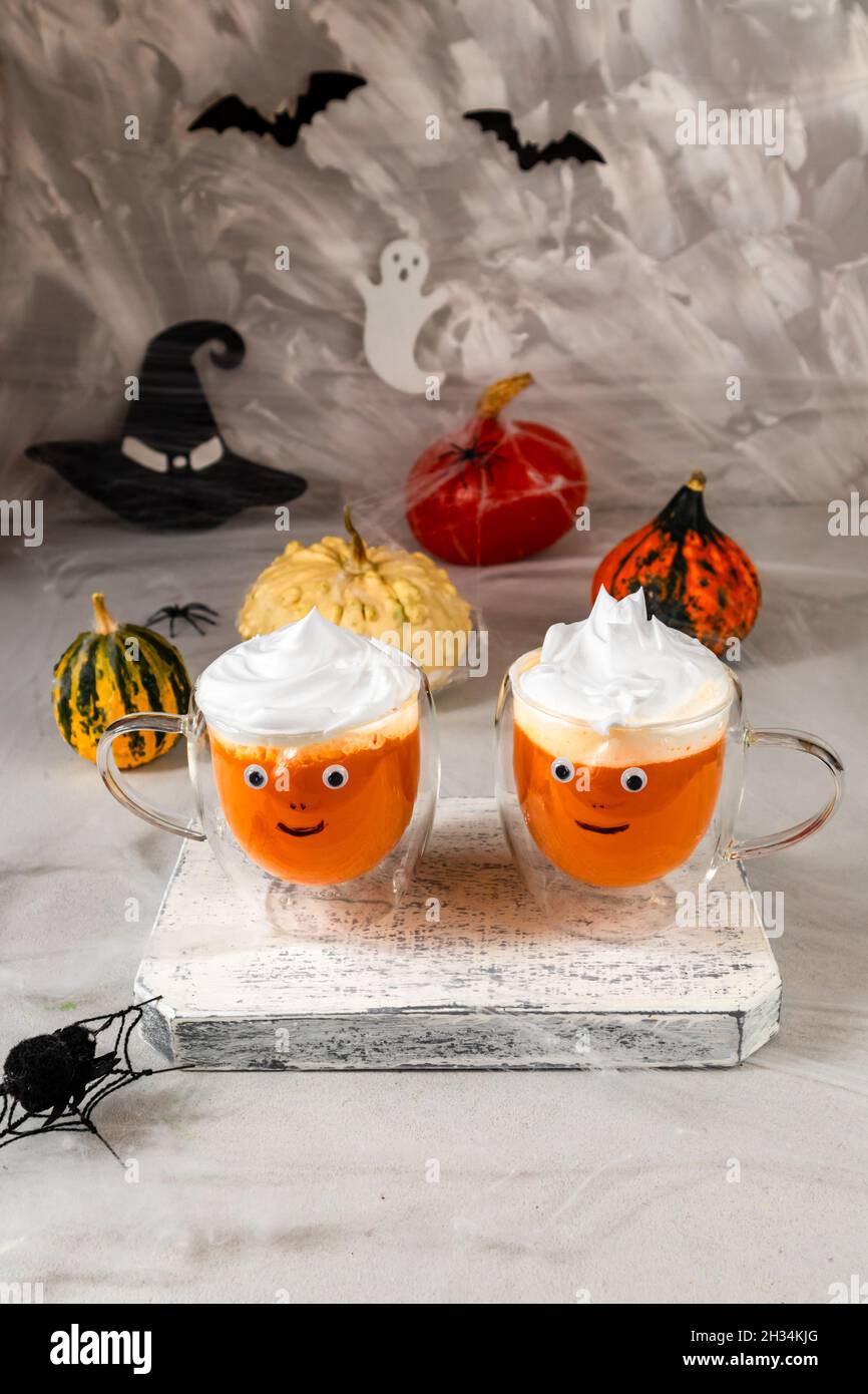 Zwei Tassen Kürbissaft aus Glas mit bemalten Gesichtern auf dem Hintergrund von Halloween-Kürbissen an einer grauen Wand mit Geistern, Fledermäusen, Spinnen, Hexenhüten Stockfoto