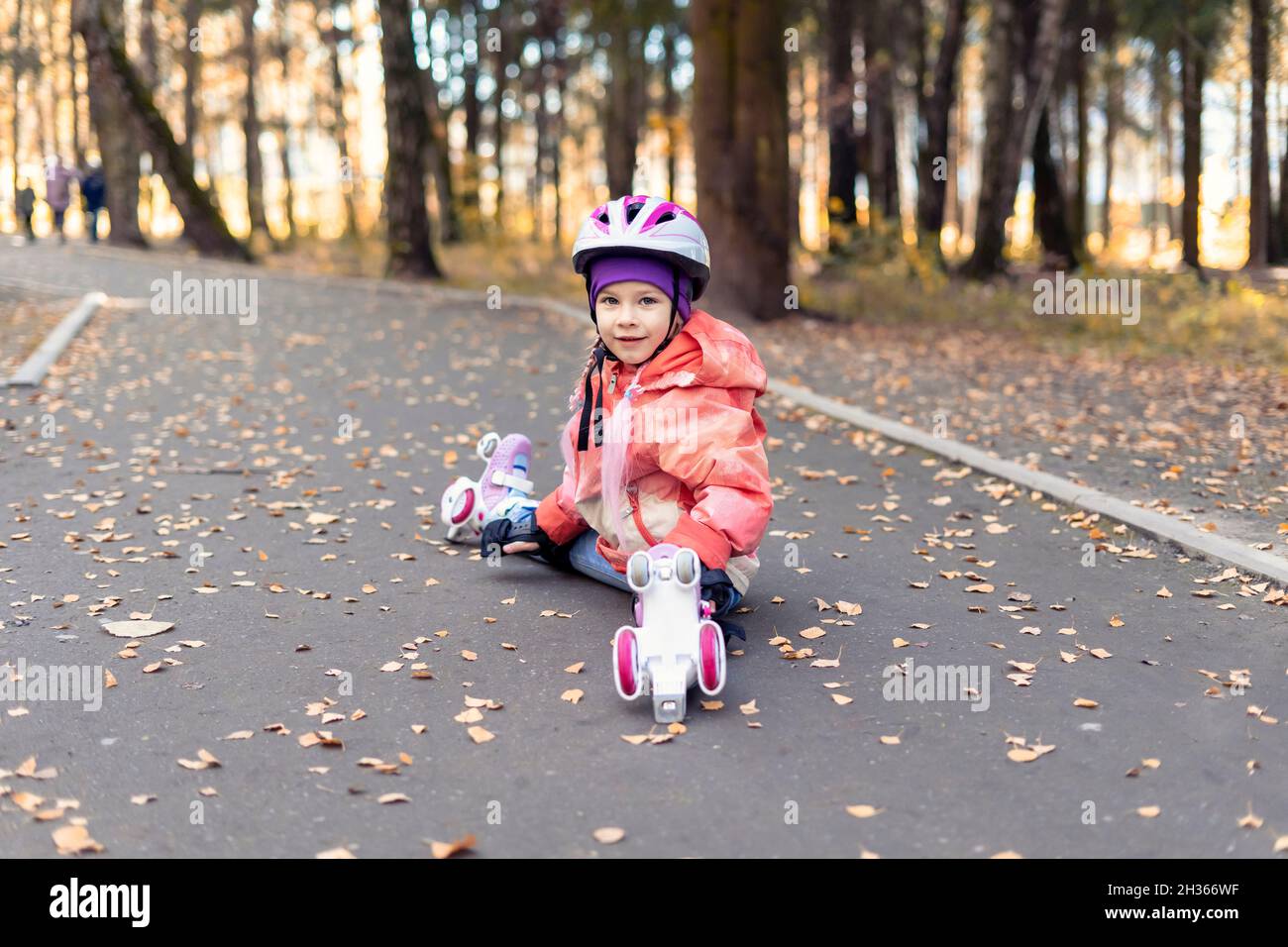 Kindermädchen in einem rosa Helm und vier Rädern Rollen sitzt auf dem Asphalt und lächelt. Herbst Outdoor-Aktivitäten Stockfoto