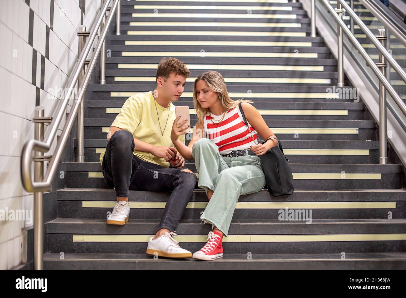 Ein junger Mann und eine junge Frau, die auf der Treppe einer U-Bahnstation sitzen. Das Mädchen zeigt dem Jungen etwas auf ihrem Handy Stockfoto