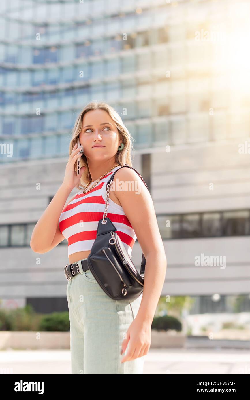 Junge hübsche blonde Frau mit Handtasche, die die Straße entlang geht und am Telefon spricht Stockfoto