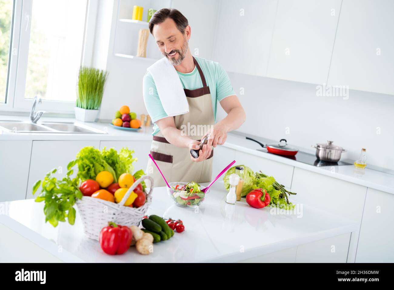 Foto von fröhlichen reifen Mann mischen Schüssel Salat halten Pfefferbox tragen Schürze blau T-Shirt Hausküche drinnen Stockfoto