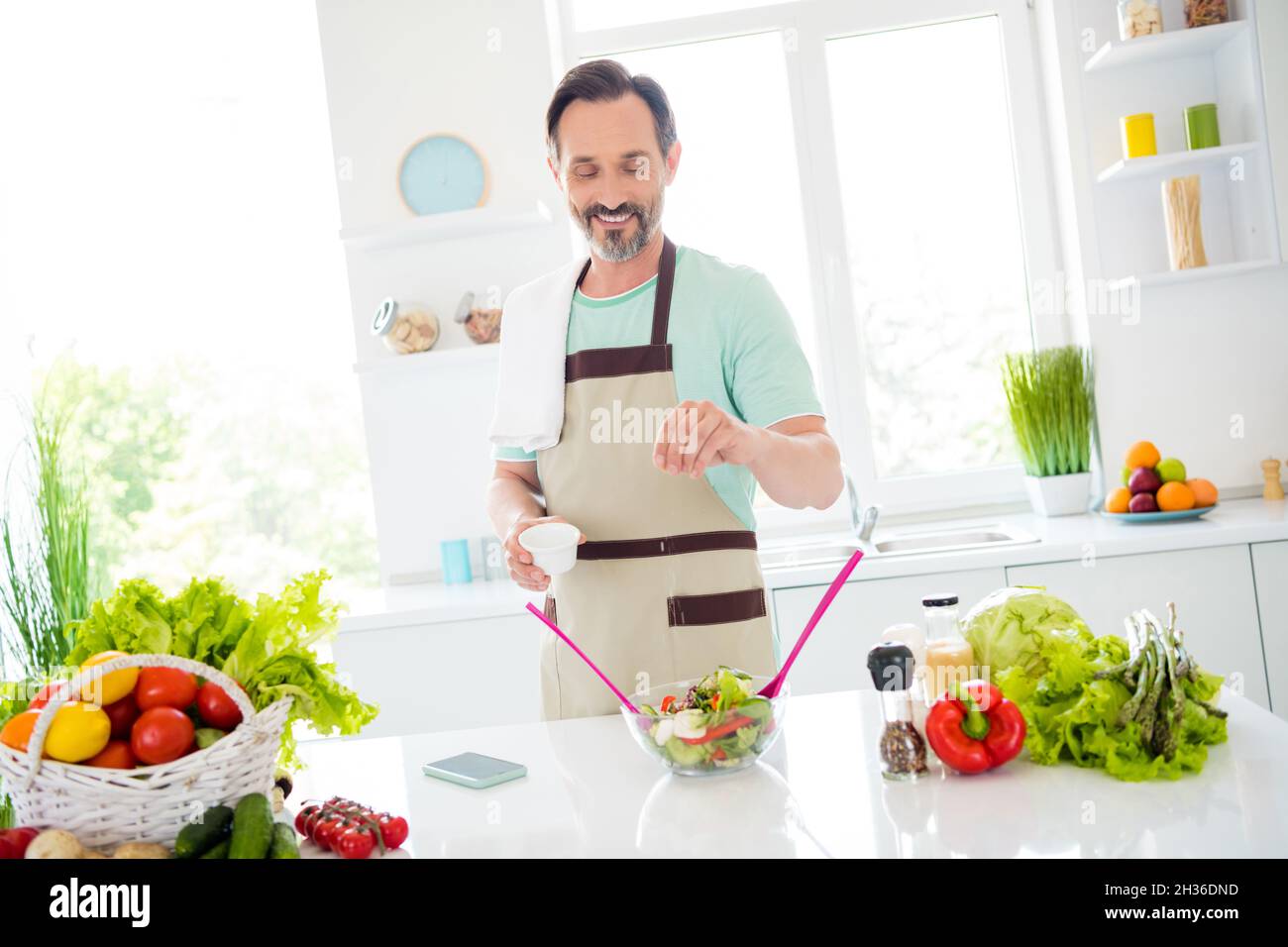 Foto von positiven Mann halten Salzbox Gewürz gemischten Salat tragen Küchengeschirr Schürze in Küche Wohnung drinnen Stockfoto