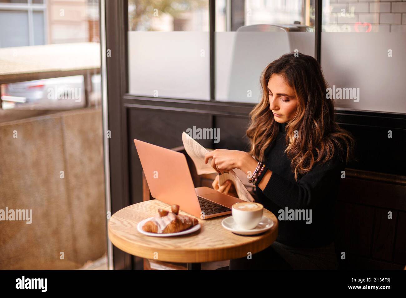 Junge Geschäftsfrau liest morgens Zeitung im Café, freiberufliche Mitarbeiterin trinkt Kaffee und genießt das Frühstück, während sie mit offenem Kaffee am Tisch sitzt Stockfoto