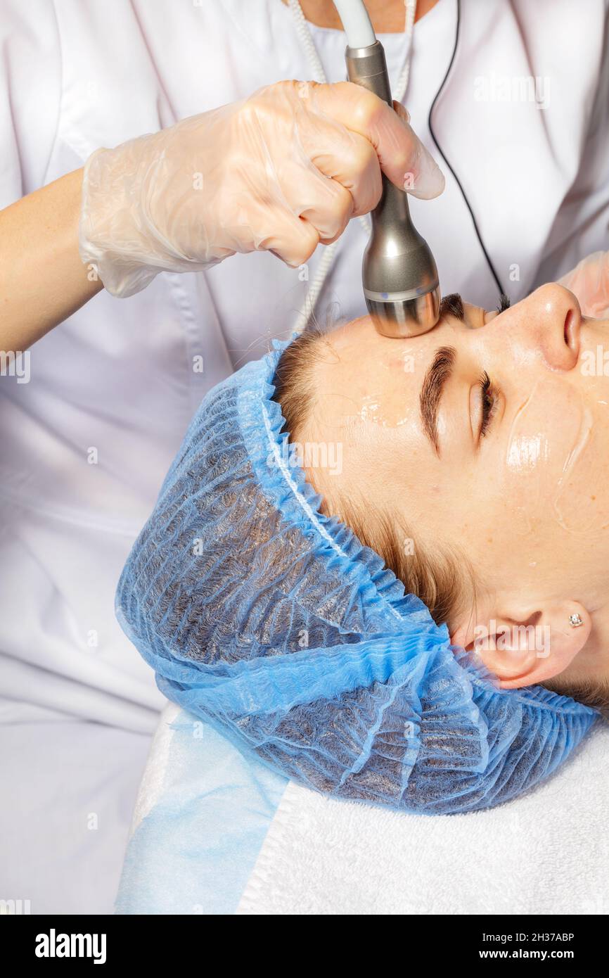 Die Kosmetikerin führt eine Sitzung der Gesichtshautmassage mit einem medizinischen Ultraschallgerät durch. Stockfoto