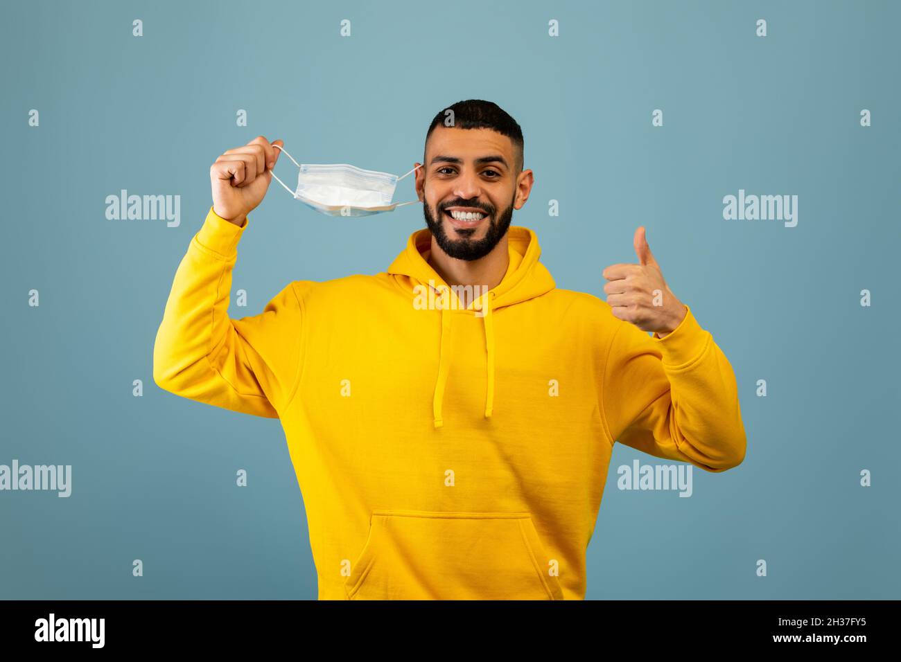 Coronavirus ist vorbei. Glücklicher arabischer Mann, der die medizinische Maske abnahm und den Daumen nach oben deuchte und über blauem Hintergrund lächelte Stockfoto