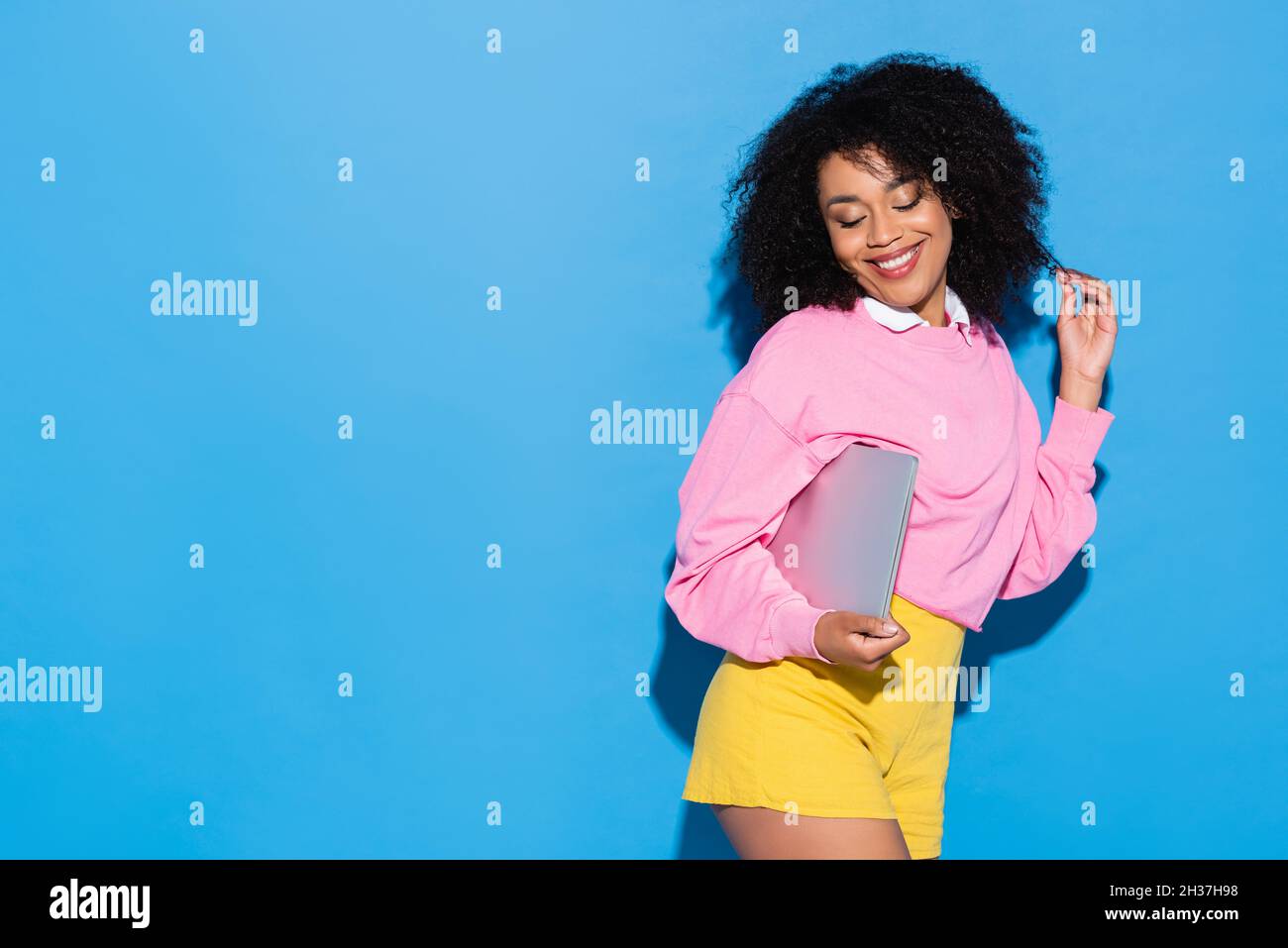 Lächelnde und flirtige afroamerikanische Frau, die mit einem Laptop auf Blau posiert Stockfoto