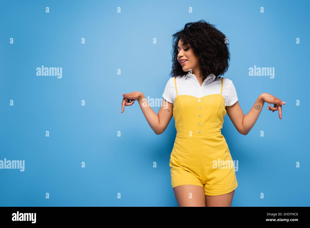 Lächelnde afroamerikanische Frau im trendigen Jumpsuit, der mit den Fingern auf Blau nach unten zeigt Stockfoto