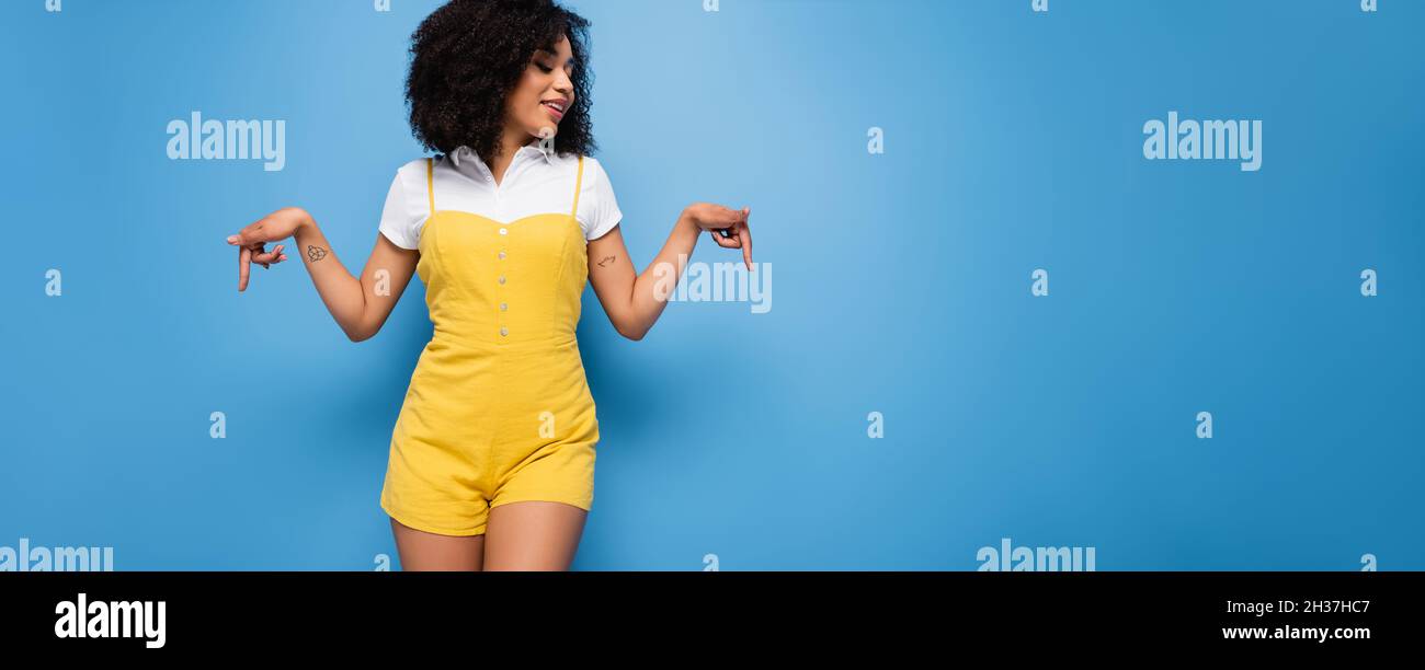 Lächelnde afroamerikanische Frau zeigt mit den Fingern auf blauem Banner nach unten Stockfoto