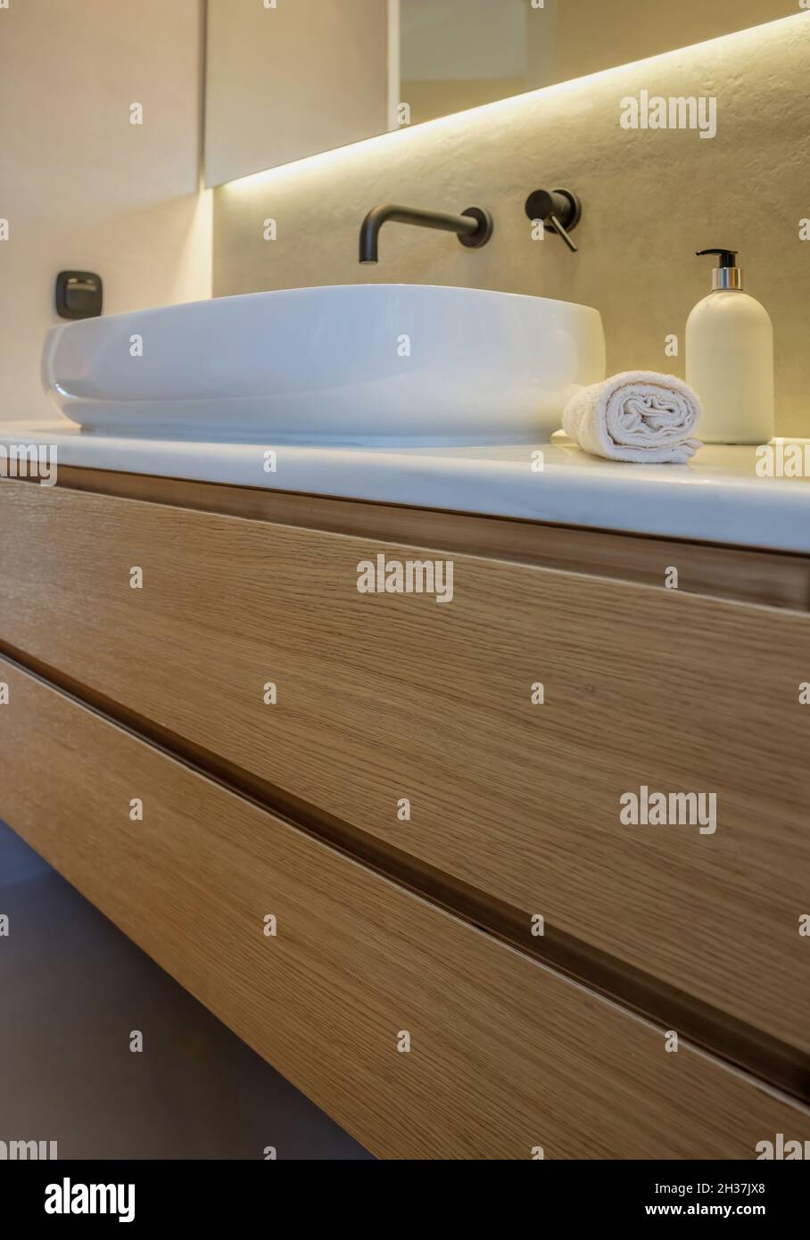 Die Badezimmereinrichtung ist modern und minimalistisch eingerichtet. Rundes Waschbecken auf weißem Marmortisch, schwarze Armaturen und Spiegel mit LED-Licht an grauer Wand, Holzaufbewahrung d Stockfoto