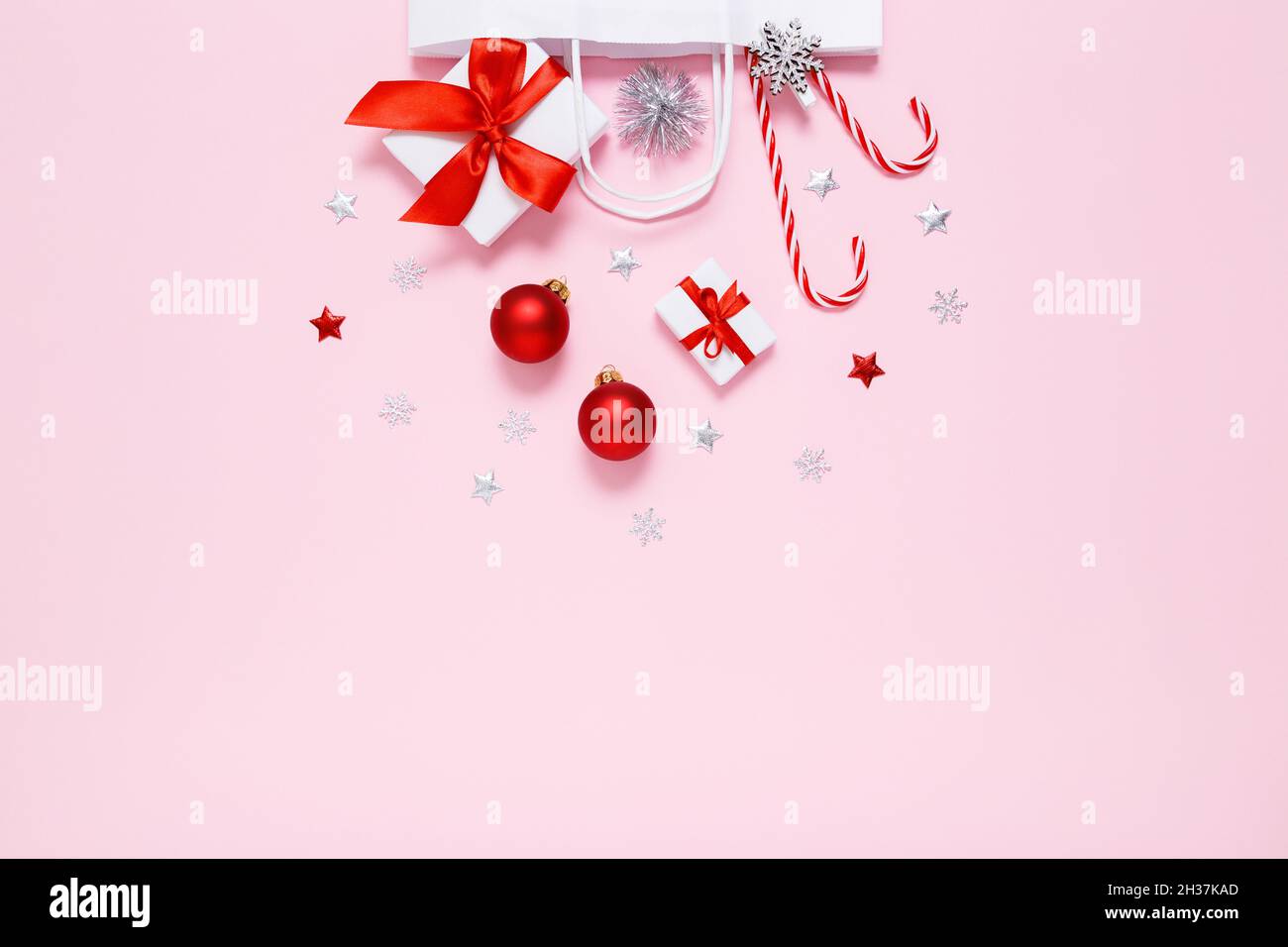 Weihnachten, Neujahr. Weiße Papiertüte mit Geschenken, süßen Bonbons, Weihnachtsdekorationen, roten Kugeln, silbernen Glitzer-Konfetti-Sternen auf pastellrosa Hintergrund. Flach liegend, Kopierplatz Stockfoto