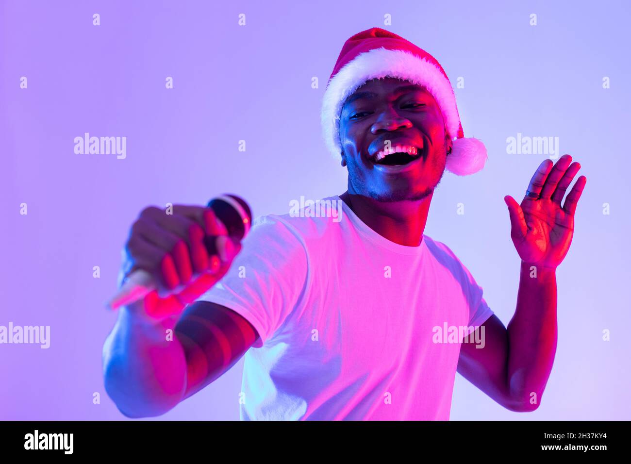 Weihnachtsmusik. Cooler Schwarzer in Santa hat singt Feiertagslied, mit Mikrofon, Karaoke in Neonlicht Stockfoto