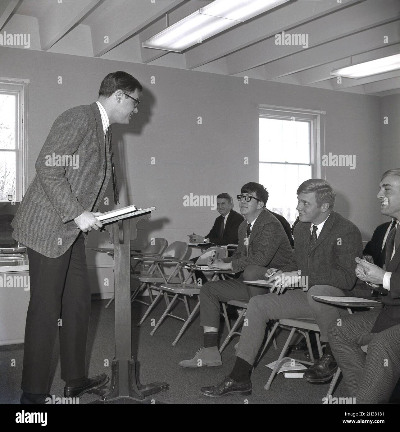 1960er Jahre, historisch, in einem Klassenzimmer an einer Geschäftshochschule, ein erwachsener männlicher Dozent, der an einem hölzernen Rednerpult steht und mit einer Gruppe gut gekleideter männlicher Studenten spricht, die an Metallstühlen sitzen, möglicherweise Postgraduierte, die anscheinend seinen Unterricht in Virginia, USA, genießen Stockfoto