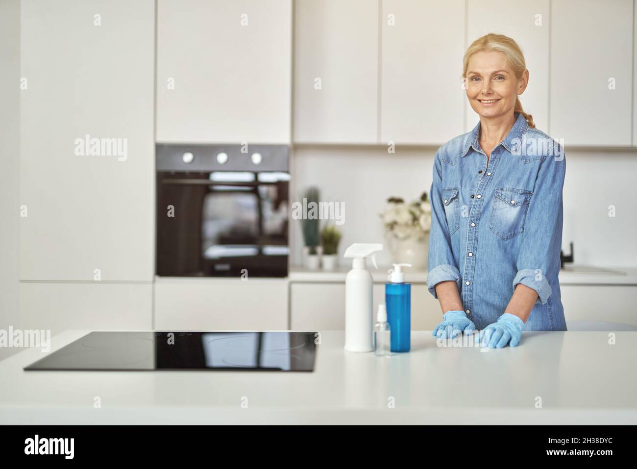 Lächelnde Frau in Gummihandschuhen, bereit zum Aufräumen in der Küche. Reinigungsmittel, Desinfektionsmittel und Gel in der Flasche auf der Oberfläche Stockfoto