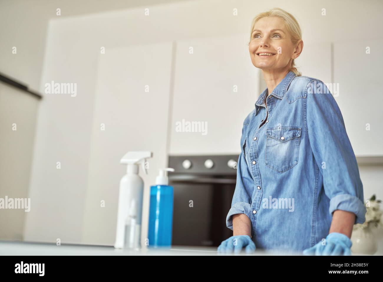 Attraktive reife Frau in Gummihandschuhen lächelt weg, bereit für die Reinigung in der Küche. Reinigungsmittel, Desinfektionsmittel und Gel in der Flasche auf der Oberfläche Stockfoto