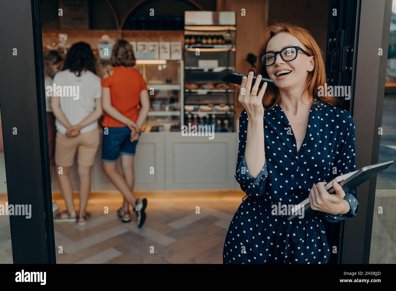 Rotschopf junge Frau macht Sprachanruf hält Smartphone in der Nähe des Mundes trägt Brille Polka Dot Kleid Stockfoto