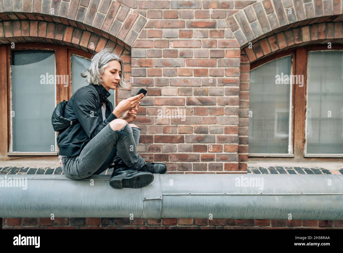 Frau sitzt mit gekreuzten Beinen in der Nähe von Backsteingebäude und schaut auf den Telefonbildschirm Stockfoto