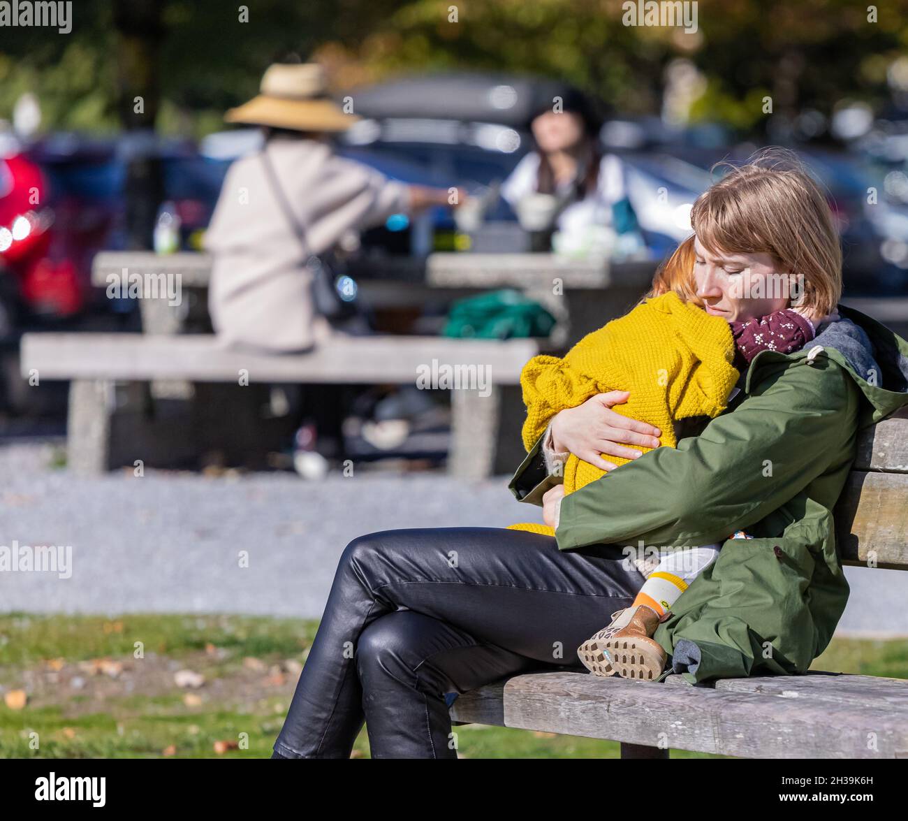 Kleine traurige Tochter umarmte ihre Mutter. Mutter umarmt ihr kleines Kind im Freien in einem Park. Herbstsaison. Blick auf die Straße, verschwommener Hintergrund, Konzeptstudie Stockfoto