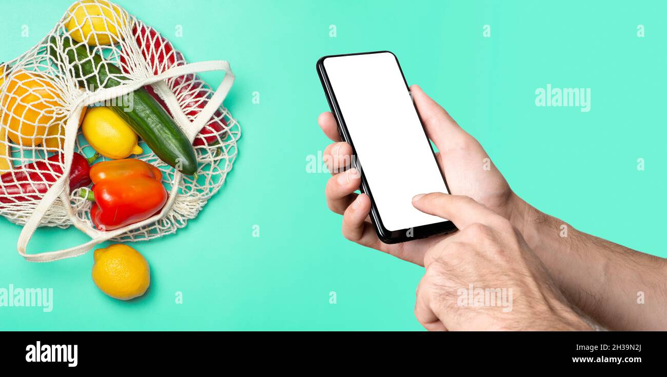 Lebensmittelgeschäft online Hintergrundkonzept. Eine Hand mit Smartphone und umweltfreundlicher Tasche mit Gemüse und Obst aus dem Supermarkt. Bestellen, Lebensmittelgeschäft und Lieferung von Lebensmitteln aus dem Öko-Markt. Hochwertige Fotos Stockfoto