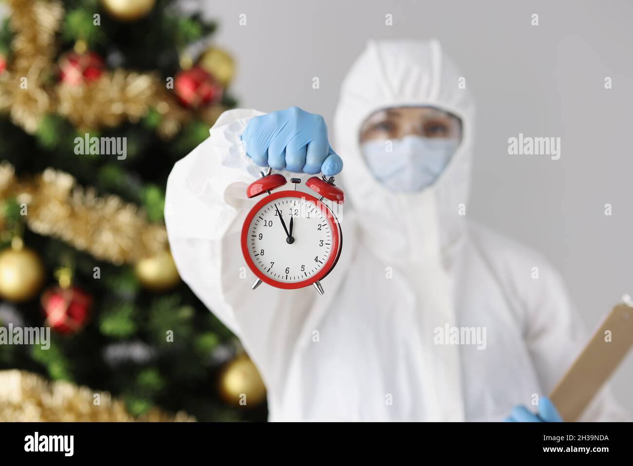 Arzt in schützendem Anti-Pest-Anzug mit Wecker in der Nähe des weihnachtsbaums Stockfoto