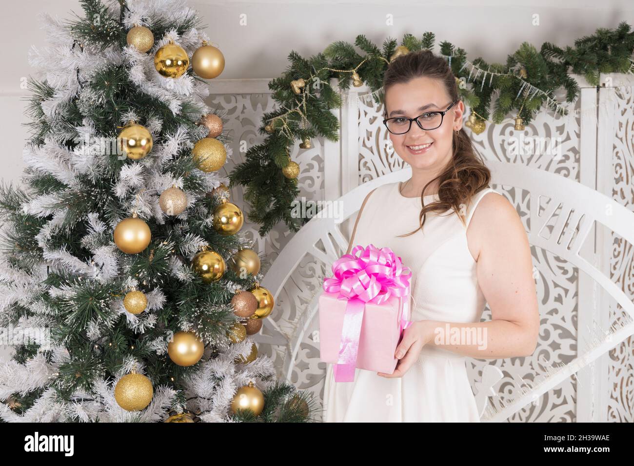 Eine junge Frau mit einem charmanten Lächeln und einer Brille hält eine Geschenkbox mit einer rosa Schleife in der Nähe des Weihnachtsbaums Stockfoto