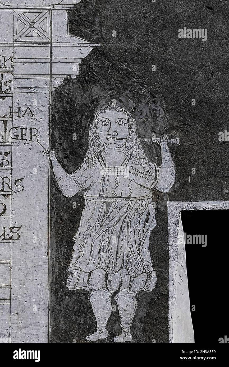 Eine Frau oder ein Mädchen in einem knielangen Kleid steht mit einem Horn neben einer Inschrift in romanischer Sprache aus dem Jahr 1664 in diesem charmanten Detail rustikaler Sgraffito-Kunst an der Fassade eines traditionellen Engadinerhauses in Ardez im Unterengadiner Tal, Kanton Graubünden oder Graubünden, Ostschweiz. Stockfoto