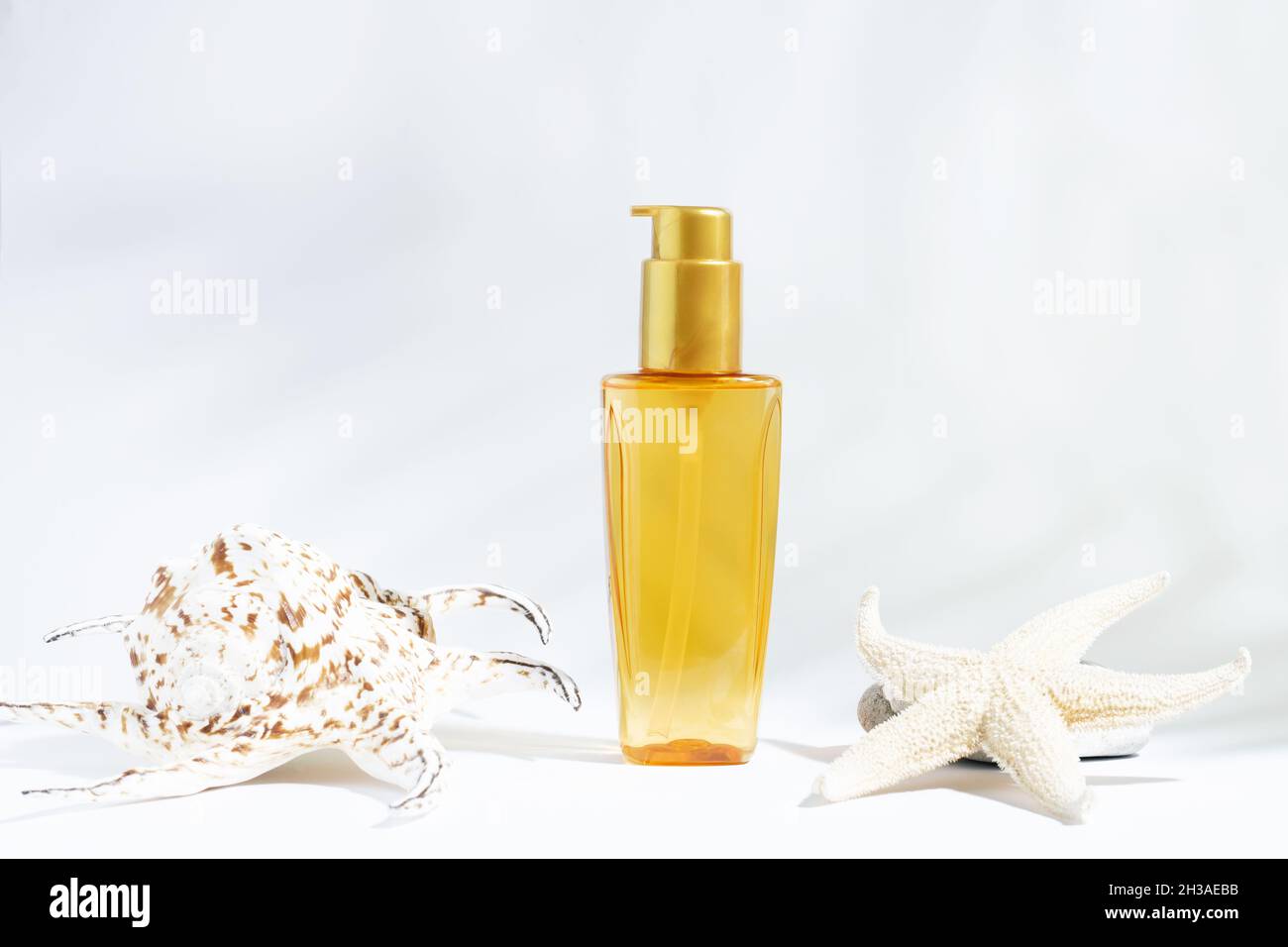 Klare Flasche gelb-orangefarbenes Öl für Sonne, Bräunung oder Haarschutz. Weißer Hintergrund mit kosmetischem Öl und Muscheln. Pflegekosmetik im Sommer. Stockfoto