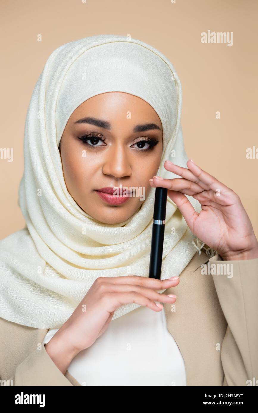 Junge muslimische Frau in Hijab hält Mascara isoliert auf beige Stockfoto