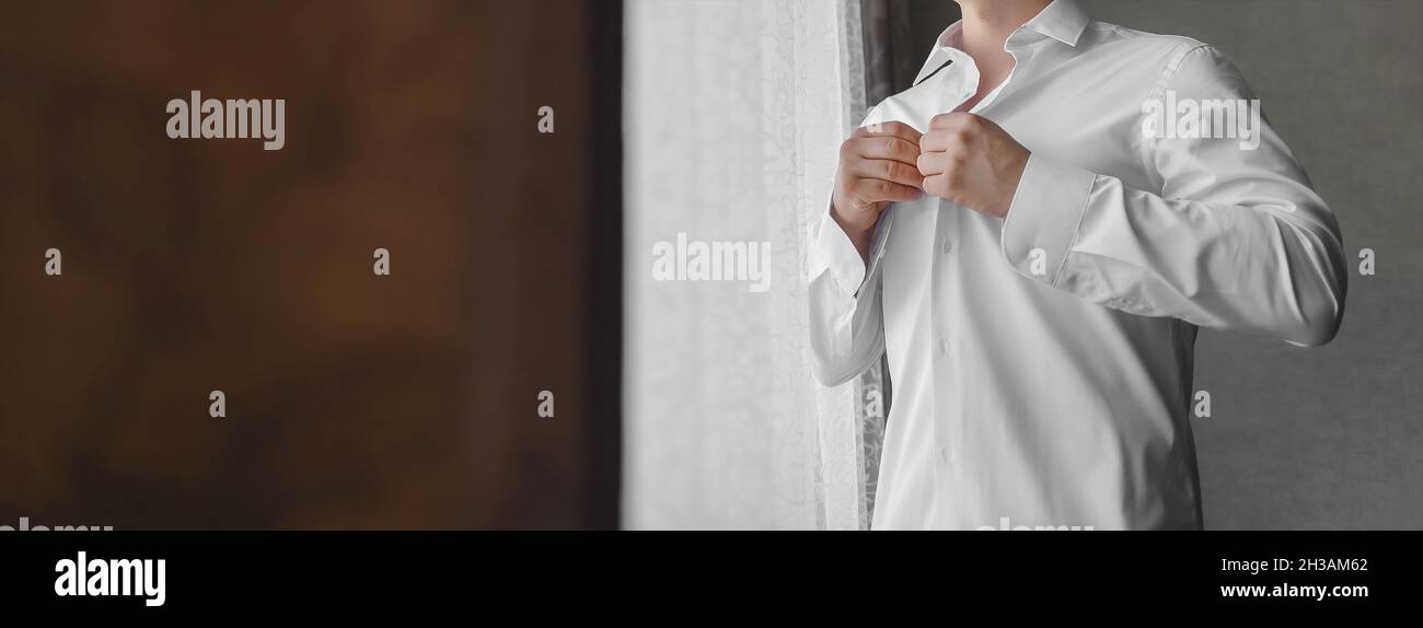 Ein Mann klammelt Knöpfe auf einem weißen Hemd mit seinen Händen im Licht vom Fenster. Stockfoto