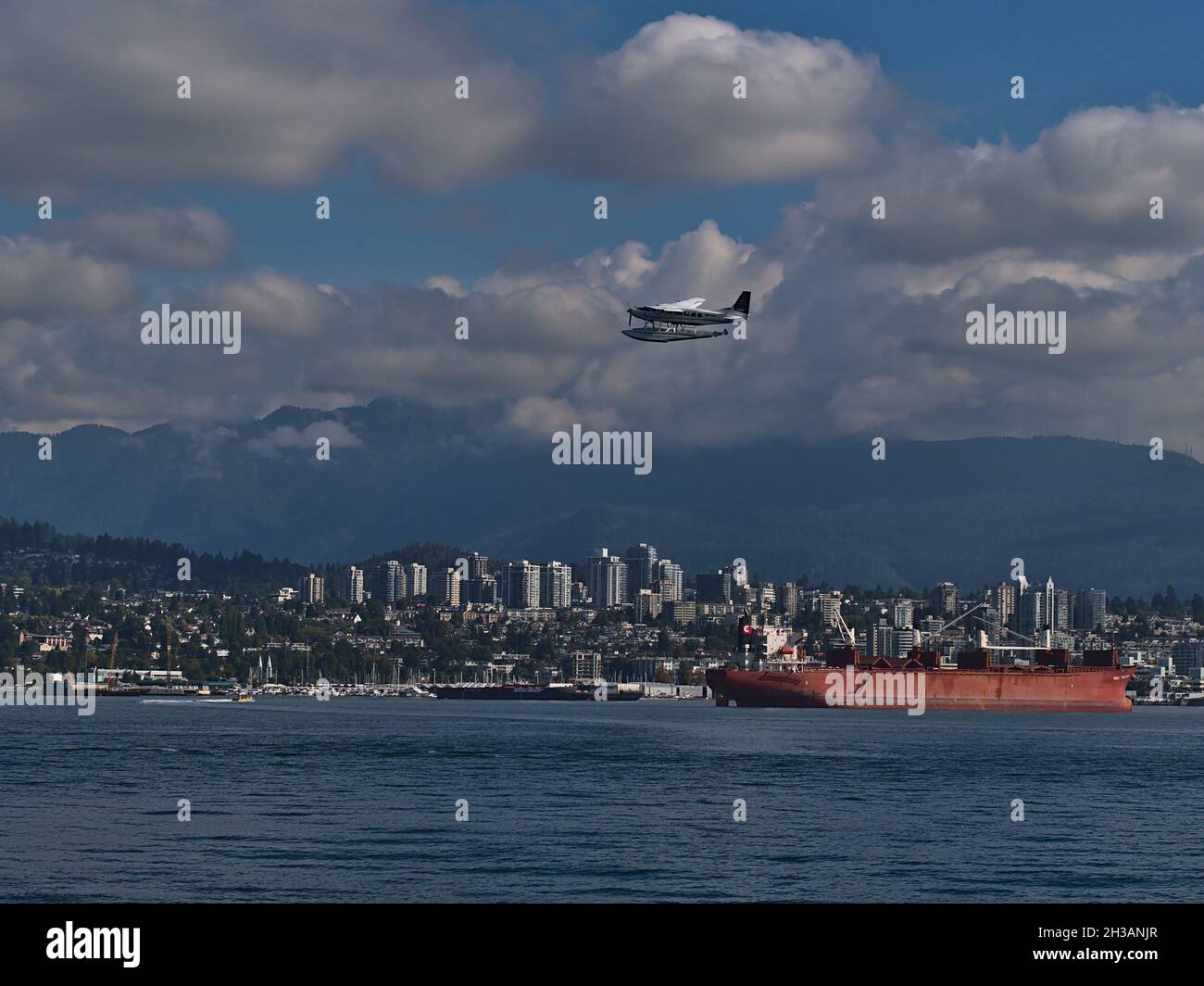 Rotes Massenfrachtschiff vor Anker im Burrard Inlet vor der Innenstadt von North Vancouver mit Wasserflugzeug, das vorbeifährt und Bergen im Hintergrund. Stockfoto