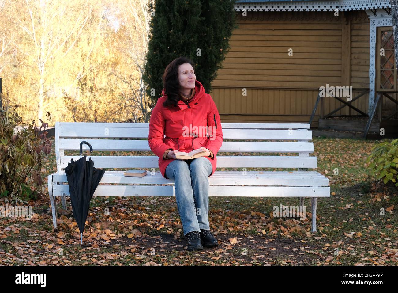 Eine Frau in einer roten Jacke sitzt auf einer weißen Bank im Park und liest Ein Papierbuch. Stockfoto