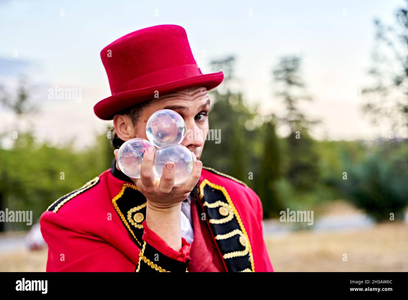 Bärtiger Mann in rotem Hut, der einen Haufen durchsichtiger Glaskugeln zeigt und während der Performance im Park auf die Kamera schaut Stockfoto