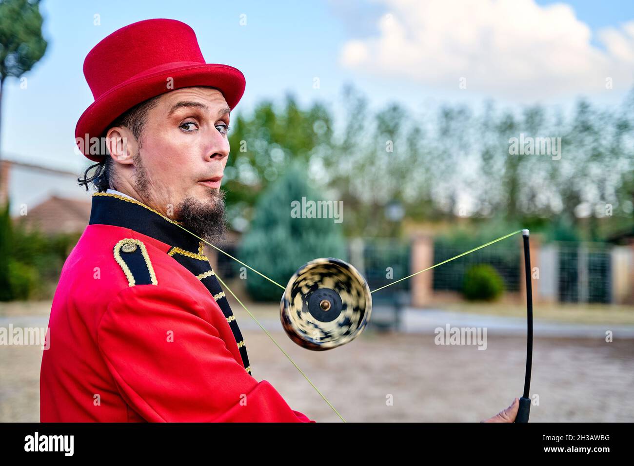 Seitenansicht eines exzentrischen bärtigen Mannes in rotem Kostüm und Hut, der mit Diabolo jongliert und die Kamera auf dem unscharfen Hintergrund des Parks anschaut Stockfoto