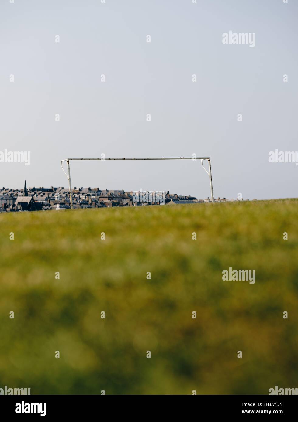 Ein einsames Fußball-/Fußballtor ohne Netz in einem leeren Sommerrasenfeld mit der entfernten Stadt Whitby und Gebäuden dahinter. Stockfoto