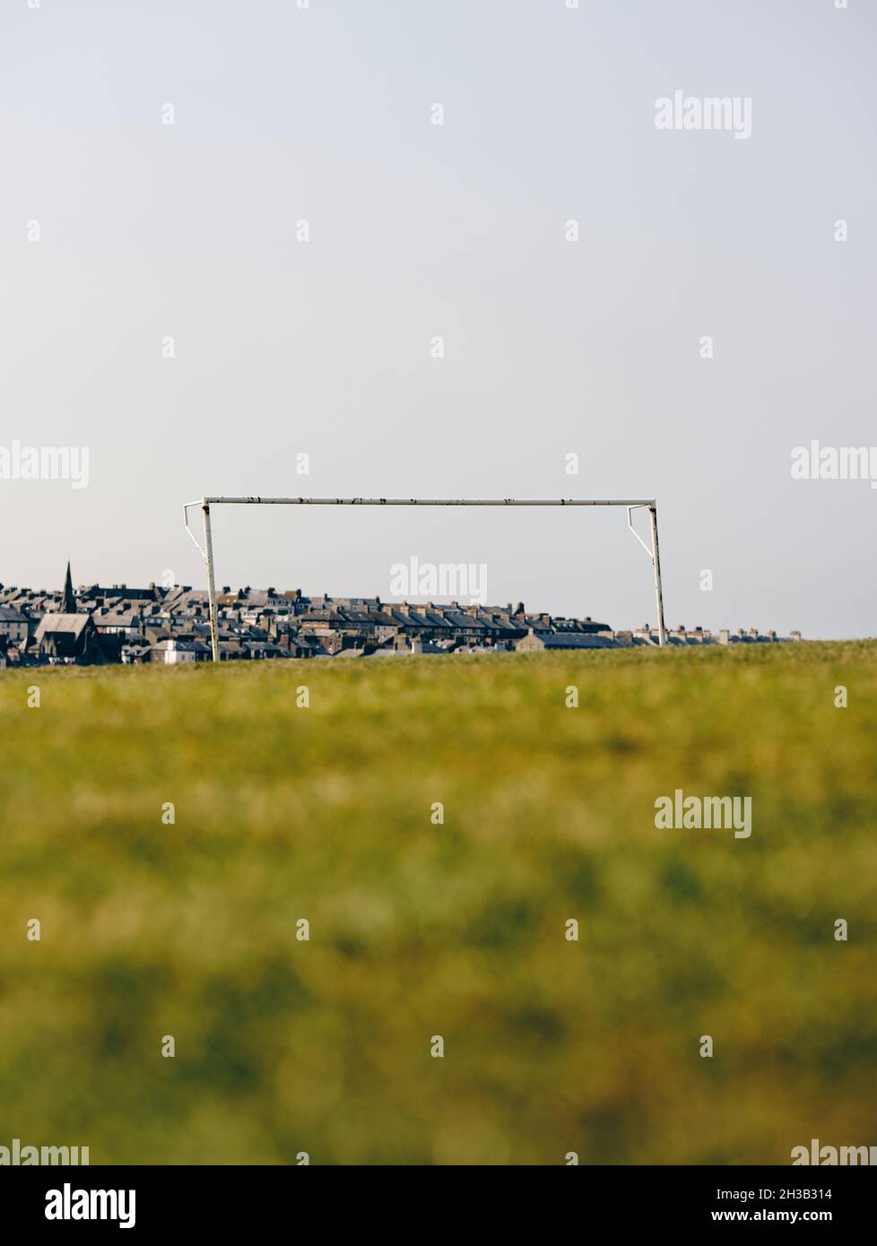 Ein Einzelfußball-/Fußballtor ohne Netz in einem leeren Sommerrasenfeld mit einer unbestimmten Stadt und Gebäuden im Hintergrund. Stockfoto