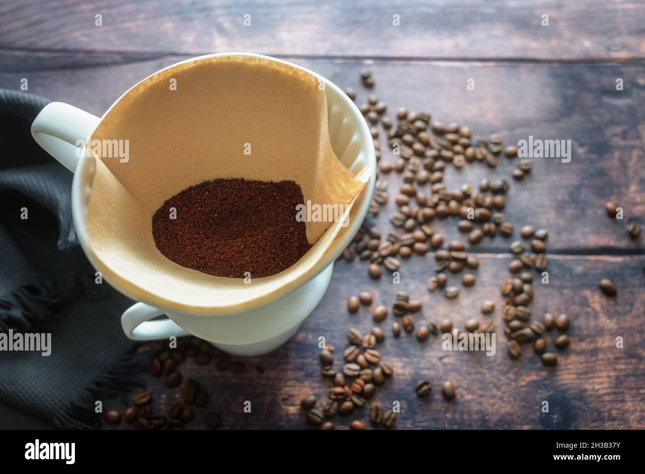 Gemahlener Kaffee in einem Papierfilterbeutel und in einem Porzellanhalter auf einem Becher für ein aromatisch tropfendes Heißgetränk, rustikaler Holztisch mit einigen Bohnen, Polizist Stockfoto