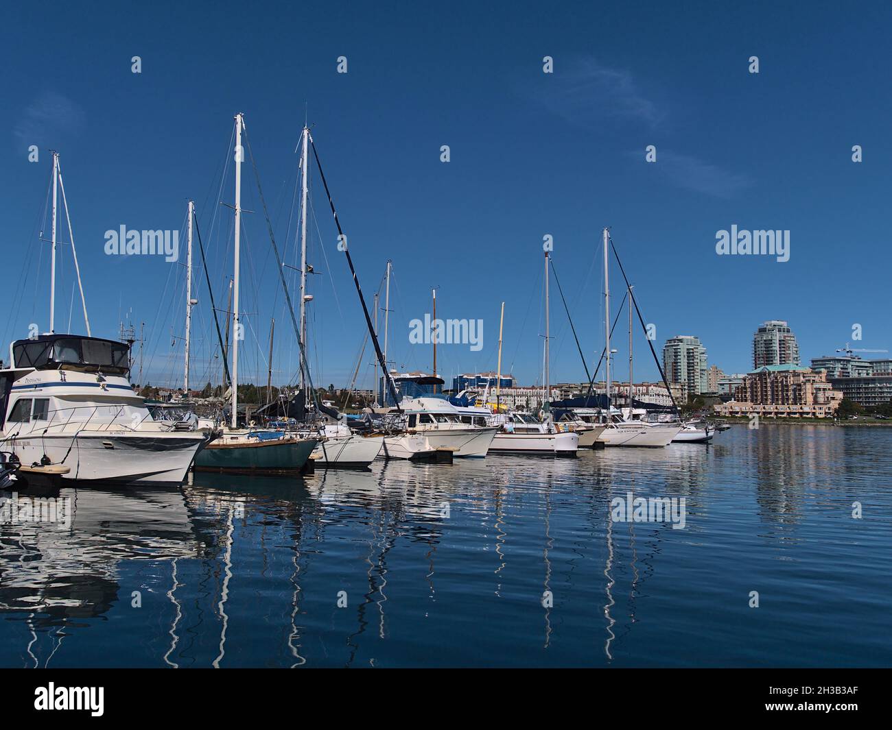 Blick auf den Yachthafen mit den Anlegebooten, die sich an sonnigen Tagen auf der glatten Wasseroberfläche von Victorias Binnenhafen mit Skyline im Hintergrund spiegeln. Stockfoto