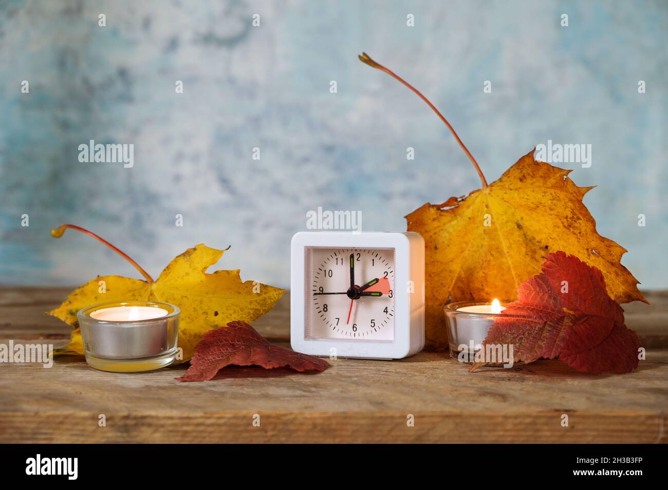Wechsel von Sommer- auf Winterzeit im Oktober, kleiner Wecker eine Stunde zurückgestellt, Herbstblätter und Kerzen auf rustikalem Holz, hellblauer Hintergrund, Stockfoto