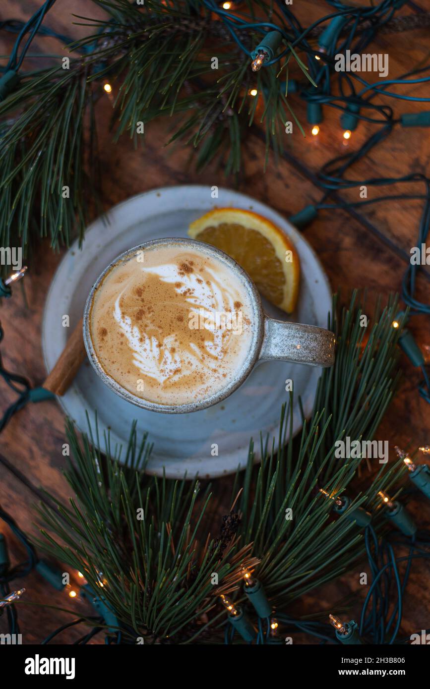 Urlaub gewürzte Zitrusfrüchte Latte mit Latte Kunst in handgefertigten grauen gesprenkelten Keramik Becher, umgeben von echten Kiefer Girlande und Weihnachtsbeleuchtung. Stockfoto
