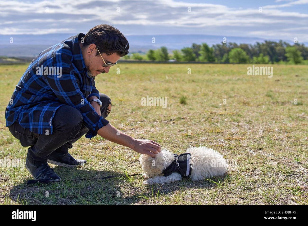 Der Latino-Mann hockte sich mit einer Sonnenbrille und streichelt seinen Pudel-Pudel-Welpen auf dem Land mit Bergen, die im Hintergrund in Argentinien nicht fokussiert waren Stockfoto