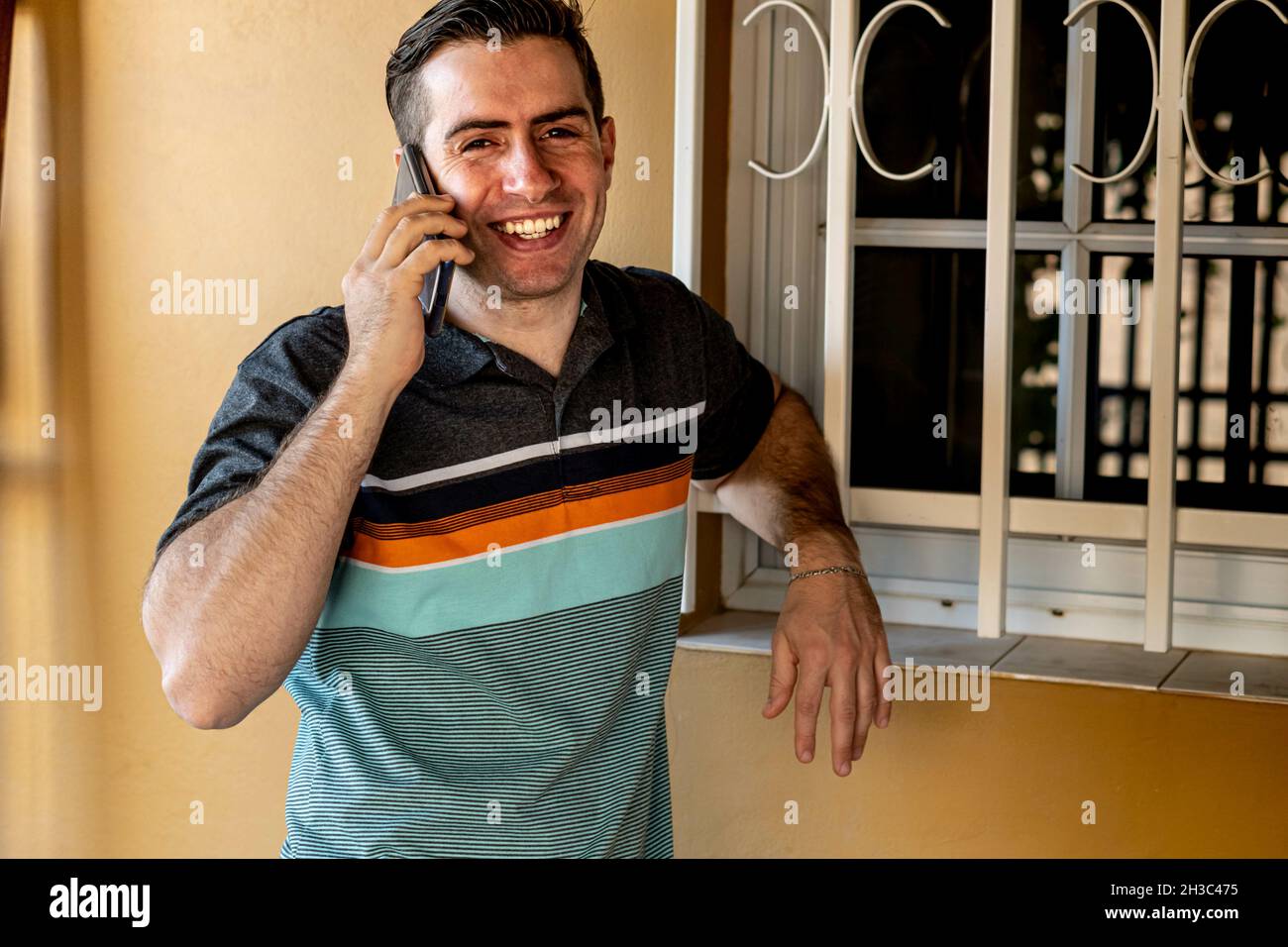Fröhlicher junger hispanischer Kerl, der sich auf seinem Telefon auf einem Fenster von draußen lehnte Stockfoto