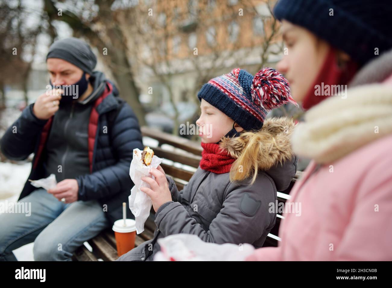 Zwei junge Schwestern und ihr Vater essen am kalten Wintertag im Stadtpark heiße Schokolade und Snacks. Papa und Kinder haben Bindungszeit. Winteraktivitäten Stockfoto
