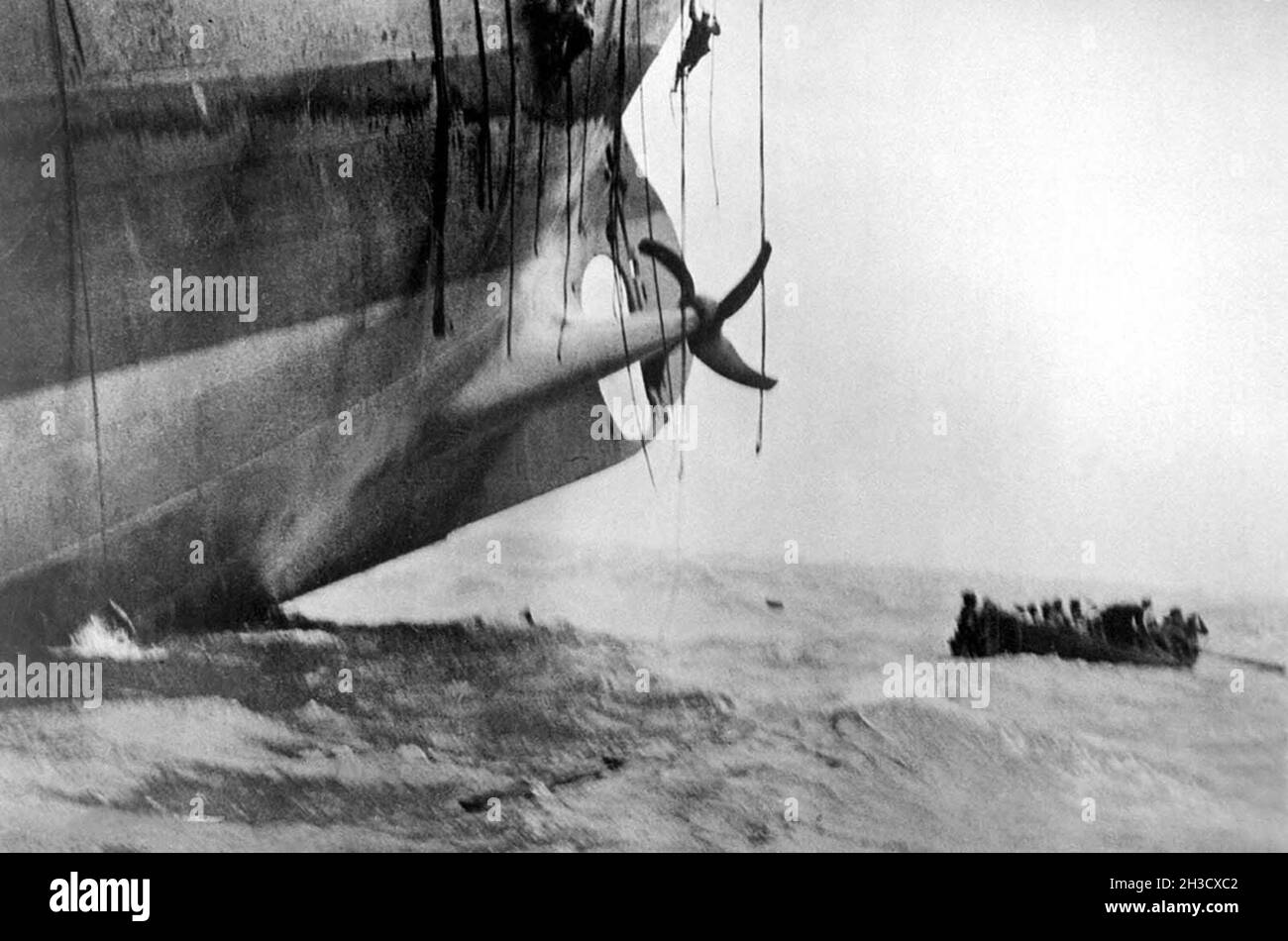 Ein dramatisches Foto eines Rettungsbootes, das sich von einem sinkenden Schiff abzieht. Das Heck des Schiffes liegt in der Luft und die letzten Besatzungsmitglieder können gesehen werden, wie sie die Seile herabsteigen. Stockfoto