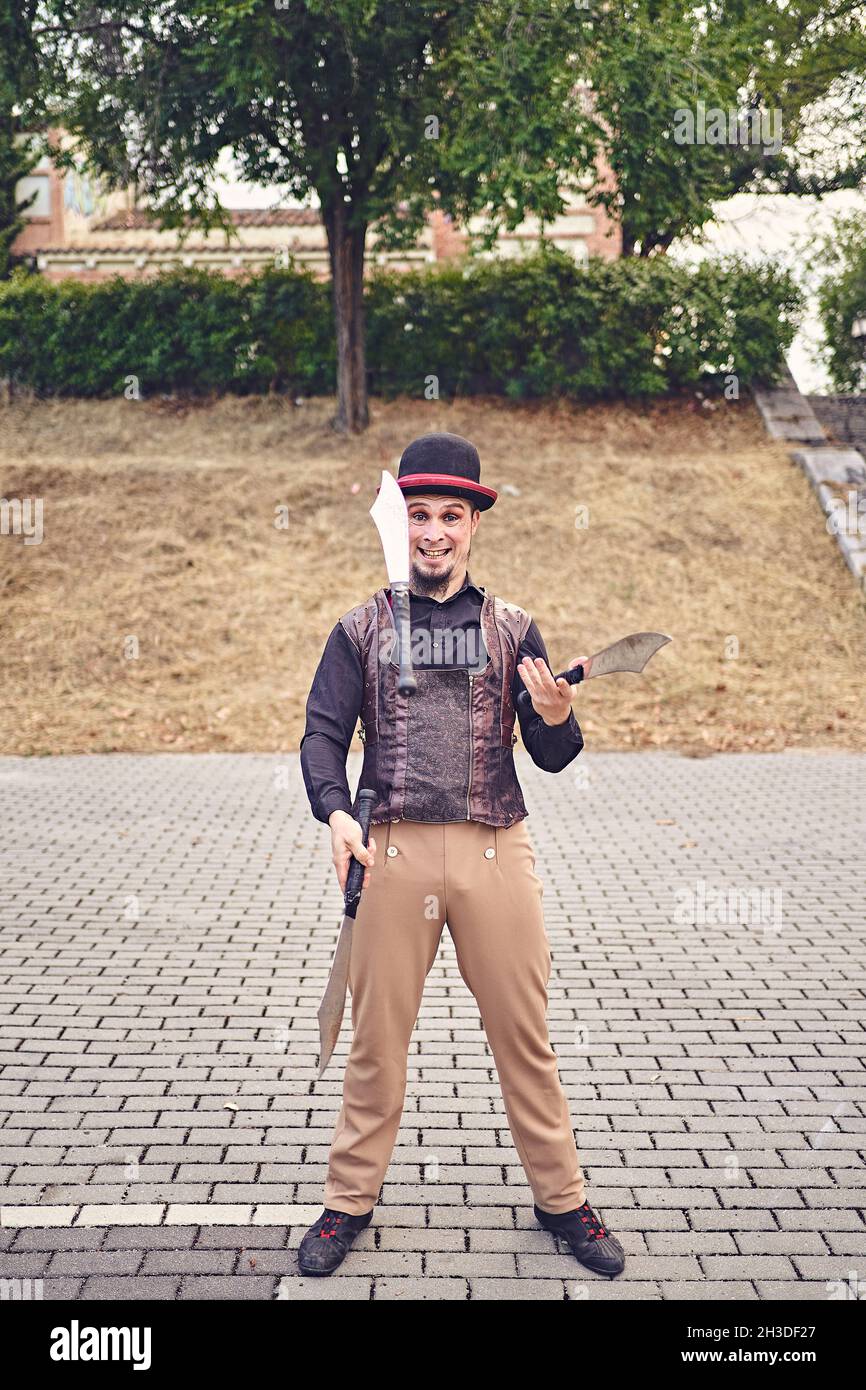Full Body lustige männliche Darsteller in Kostüm lächelnd und jonglieren Messer, während auf dem Bürgersteig während der Show im Park stehen Stockfoto
