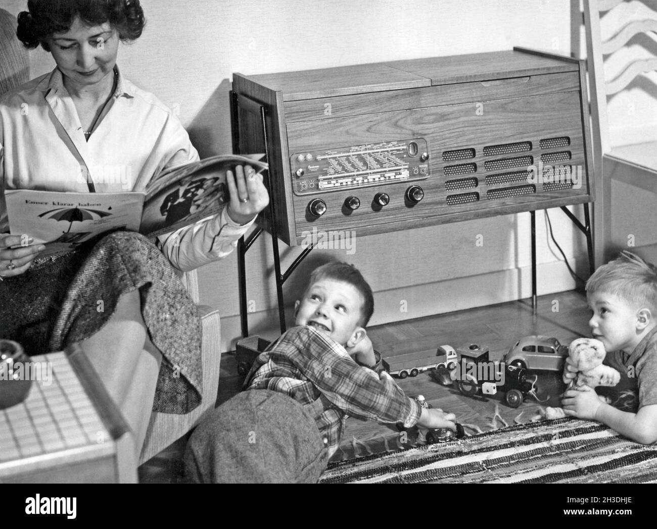 In den 1950er Jahren. Eine Mutter wird in ihrem Haus beim Lesen einer Zeitschrift gesehen, während ihre Söhne mit Spielzeugautos auf dem Boden spielen. Ein typischer Radio-Recordplayer der 1950er Jahre mit einem Holzschrank ist zu sehen. Schweden 1957 Stockfoto