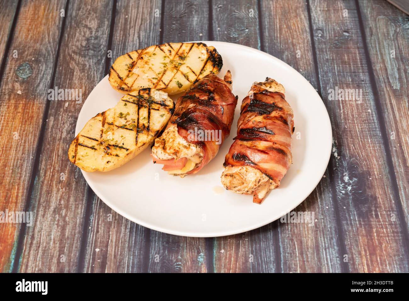 Hühnerbrötchen gefüllt mit Käse und Schinken, eingewickelt in gebratene Speckscheiben, garniert mit gegrillten Kartoffeln mit Knoblauch- und Petersiliensauce Stockfoto