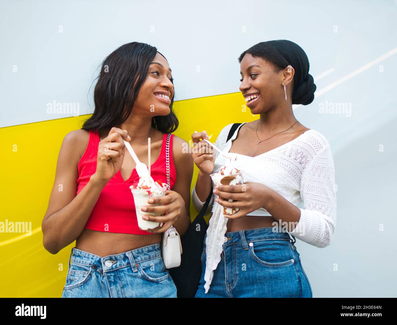 Glückliche afroamerikanische Freundinnen mit süßen Eisbechern, die sich gegenseitig angucken, während sie köstliche Desserts in der Nähe der Wand genießen Stockfoto