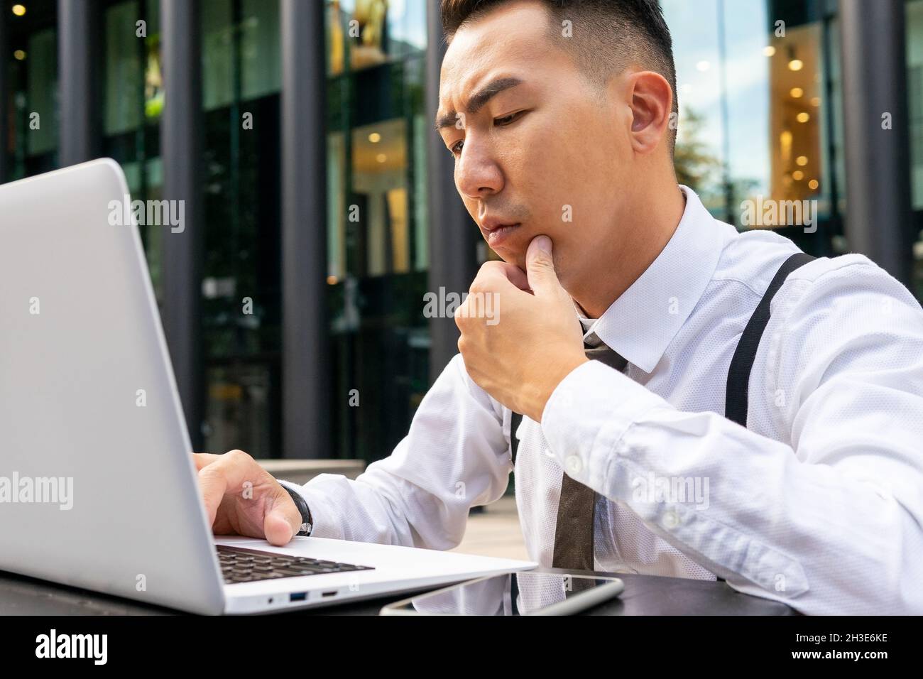 Beschneiden Sie nachdenkliche ethnische männliche Unternehmer in weißem Hemd berühren Kinn, während an Netbook an Cafeteria Tisch in der Stadt arbeiten Stockfoto