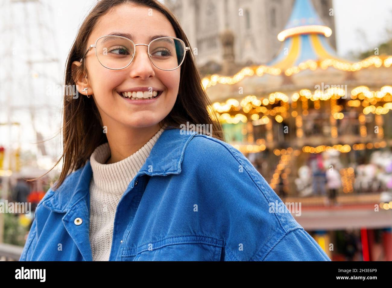 Positive junge Frau in legerer Kleidung und Brille, die bei Tageslicht in der Nähe von Gebäuden mit Karussell auf dem Hintergrund in der Stadtstraße wegschaut Stockfoto