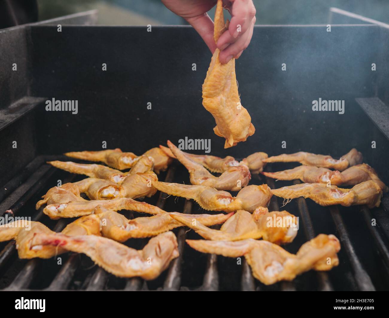 Crop anonymen Koch setzen rohe Hühnerflügel auf heiße Metall-Grill Rost mit Rauch beim Kochen auf dem Land während des Picknicks Stockfoto